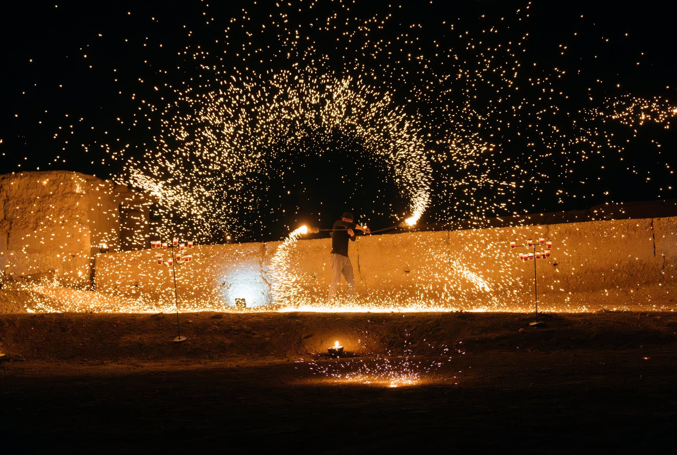 A fire performer sending showers of sparks across an earthen wall at night in Agafay